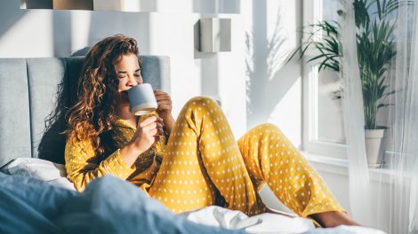 Sárga pizsamás nő ül az ágyon és kávét iszik a hálószobában