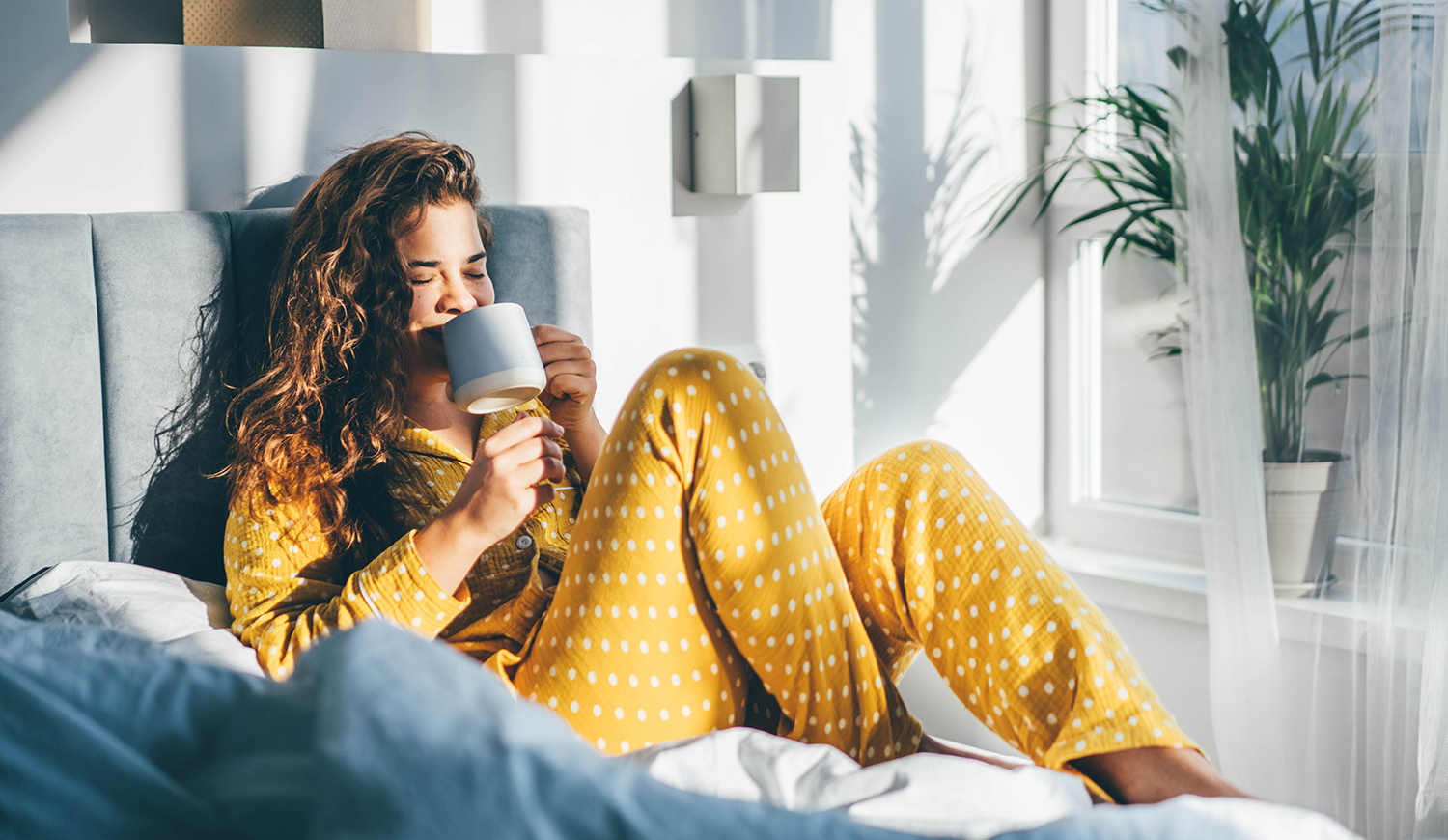 Sárga pizsamás nő ül az ágyon és kávét iszik a hálószobában