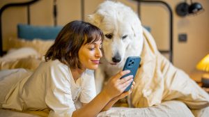 Fiatal nő okostelefont használ és egy kutyájával fekszik az ágyban