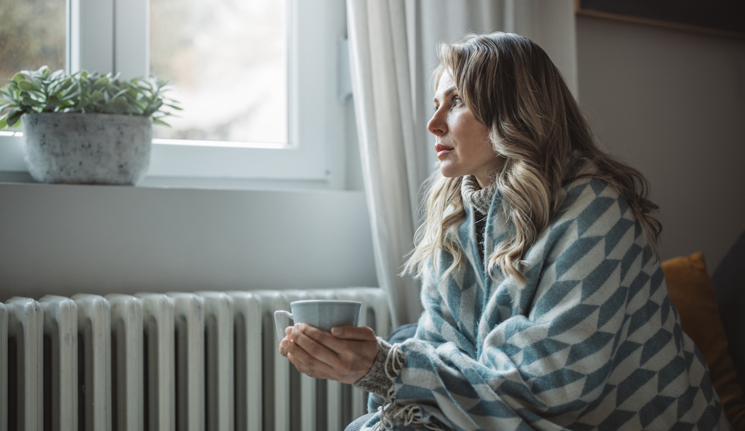 fűtés spórolás középkorú nő pokrócban ül a radiátor mellett