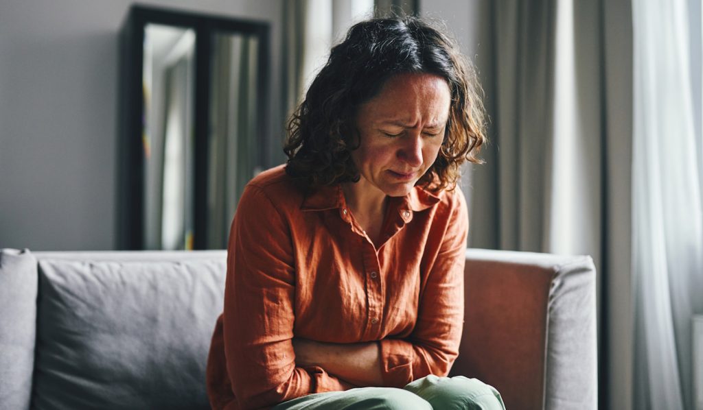 nő görnyed a kanapén, mert fáj a hasa