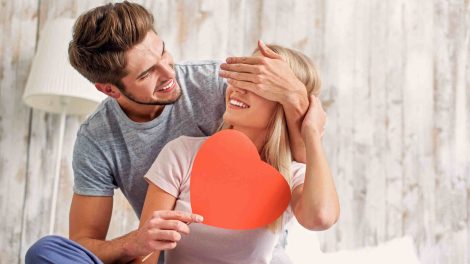 férfi játékosan befogja a nő szemét, aki papírszívet tart a kezében