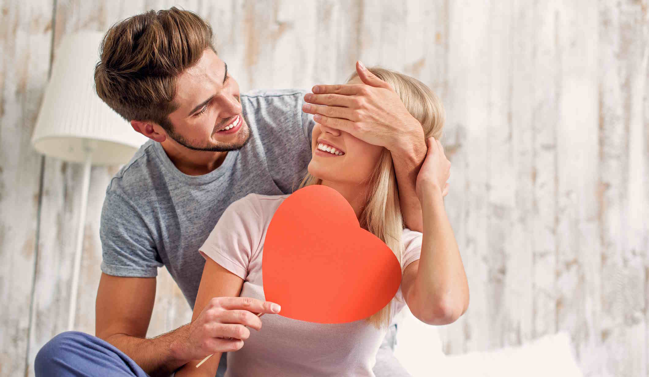 férfi játékosan befogja a nő szemét, aki papírszívet tart a kezében