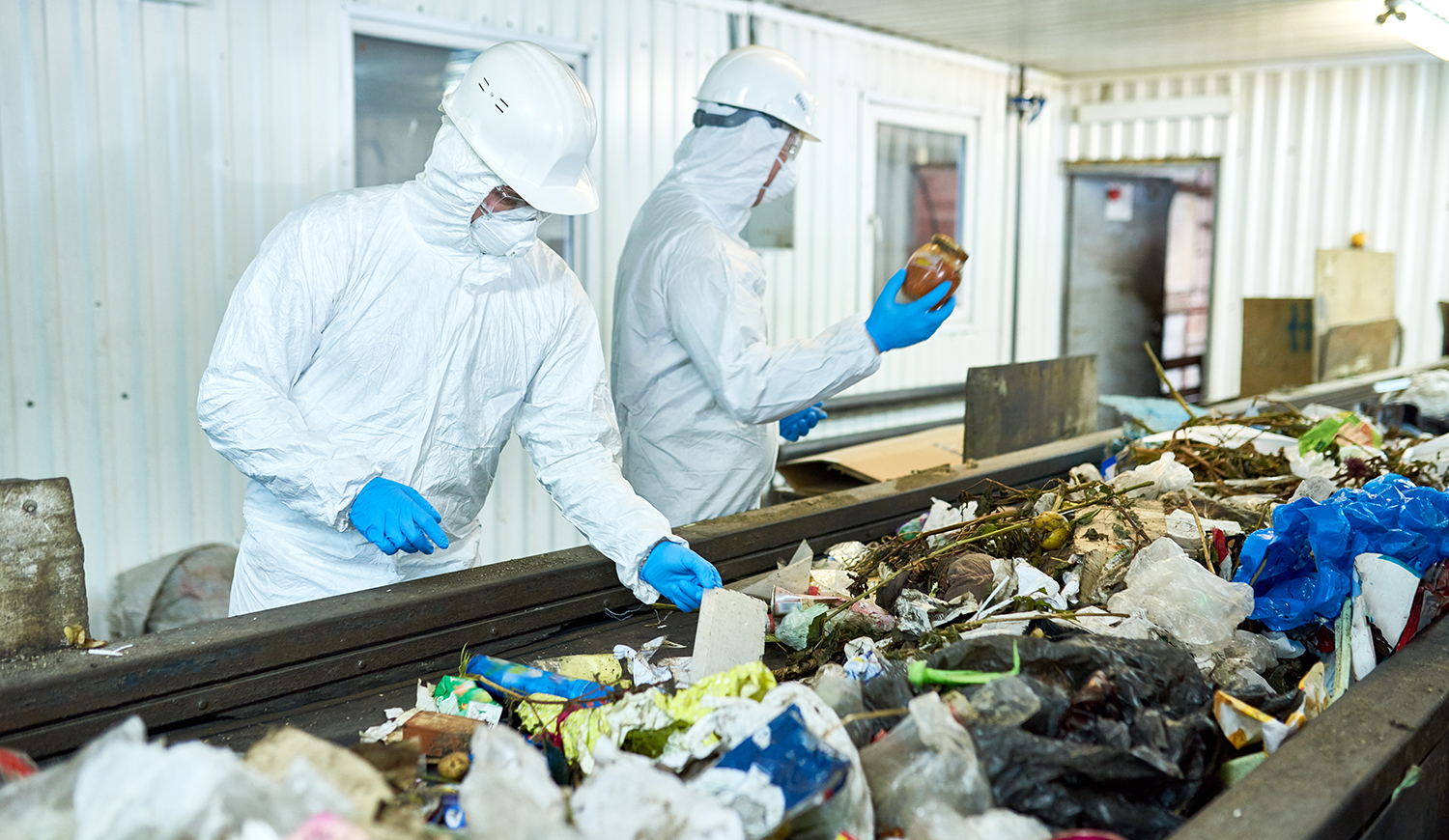 Védőruhát viselő munkás ok, akik egy hulladékfeldolgozó üzemben dolgoznak, és szállítószalagon válogatják a szemetet