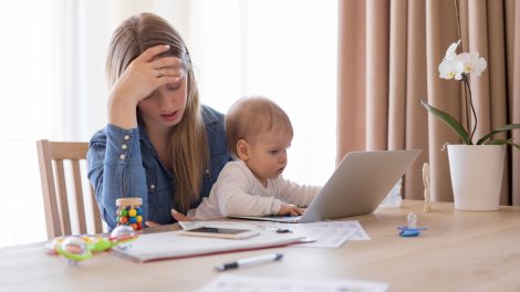 Egy anyuka ölében ül a kisgyereke, miközben próbálna dolgozni a laptopján
