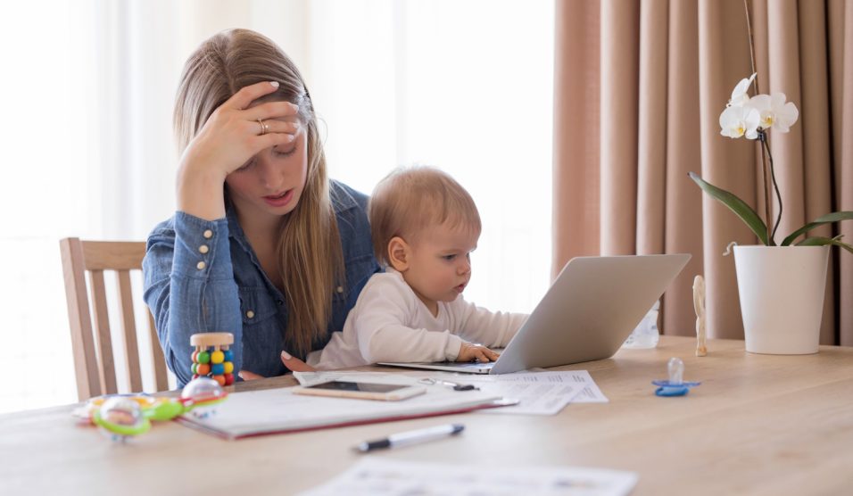 Egy anyuka ölében ül a kisgyereke, miközben próbálna dolgozni a laptopján