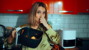 „Egy nő undorodva befogja az orrát, miközben egy szennyezett airfryer kosarat tart. A készülékben maradt ételmaradékok kellemetlen szagot árasztanak.”