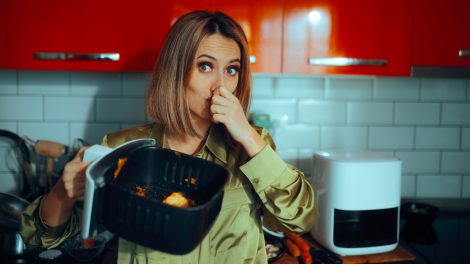 „Egy nő undorodva befogja az orrát, miközben egy szennyezett airfryer kosarat tart. A készülékben maradt ételmaradékok kellemetlen szagot árasztanak.”