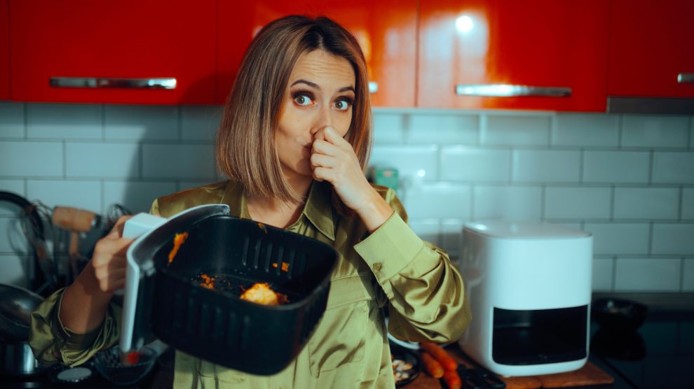 „Egy nő undorodva befogja az orrát, miközben egy szennyezett airfryer kosarat tart. A készülékben maradt ételmaradékok kellemetlen szagot árasztanak.”