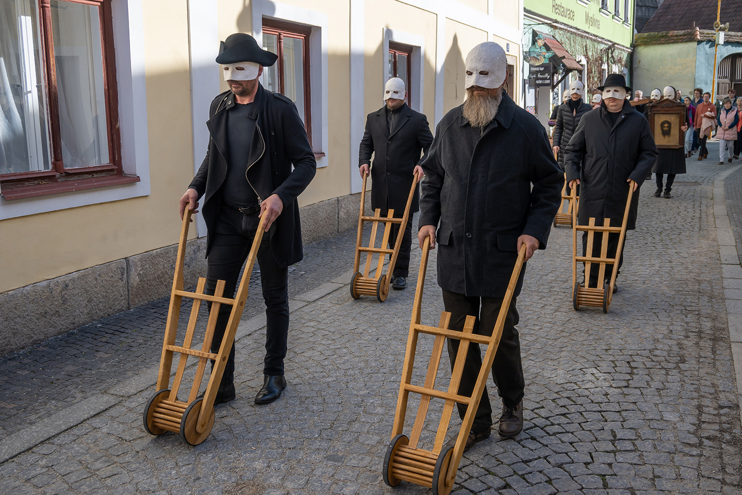 Fehér maszkot viselő emberek fa csörgőket tologatnak a nagypénteki körmenet során Trebon utcáin