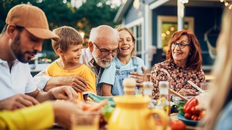 Boldog idős nagyapa az unokájával és családjával a kertben az étkezőasztalnál