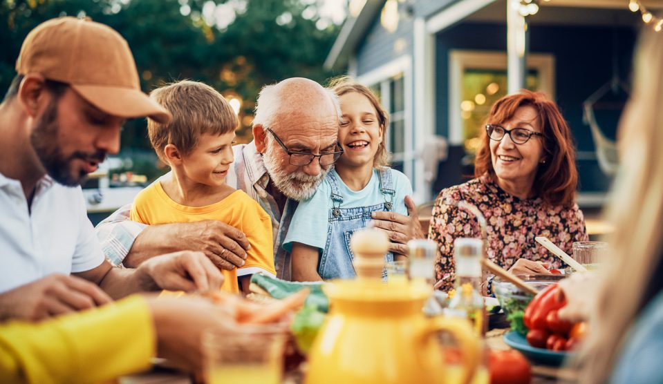 Boldog idős nagyapa az unokájával és családjával a kertben az étkezőasztalnál