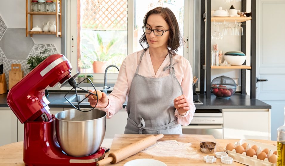 Kötényt viselő nő süteményt süt egy meghitt hangulatú konyhában. A háziasszony tojásokat üt egy karos, bolygóműves konyhai robotgép táljába. Házi készítésű sütemény vagy torta készül.