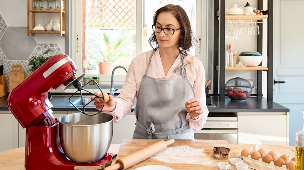 Kötényt viselő nő süteményt süt egy meghitt hangulatú konyhában. A háziasszony tojásokat üt egy karos, bolygóműves konyhai robotgép táljába. Házi készítésű sütemény vagy torta készül.