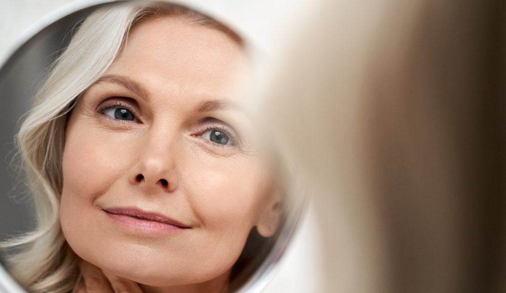 gyönyörű 50+ nő áll a tükör előtt és szépen kisminkeli magát