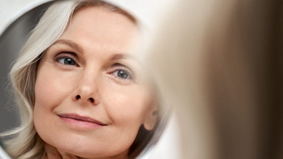 gyönyörű 50+ nő áll a tükör előtt és szépen kisminkeli magát