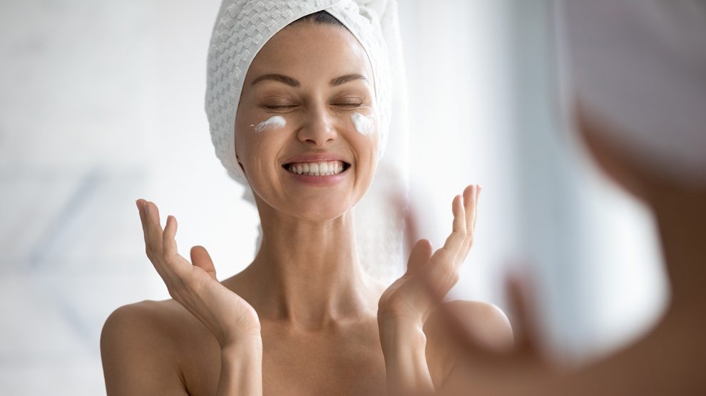 Boldog fiatal nő csukott szemmel krémezi az arcát