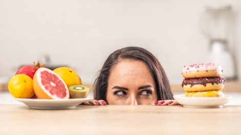 fogyni akaró nő nézi a fánkokat miközben van mellette egy tál gyümölcs