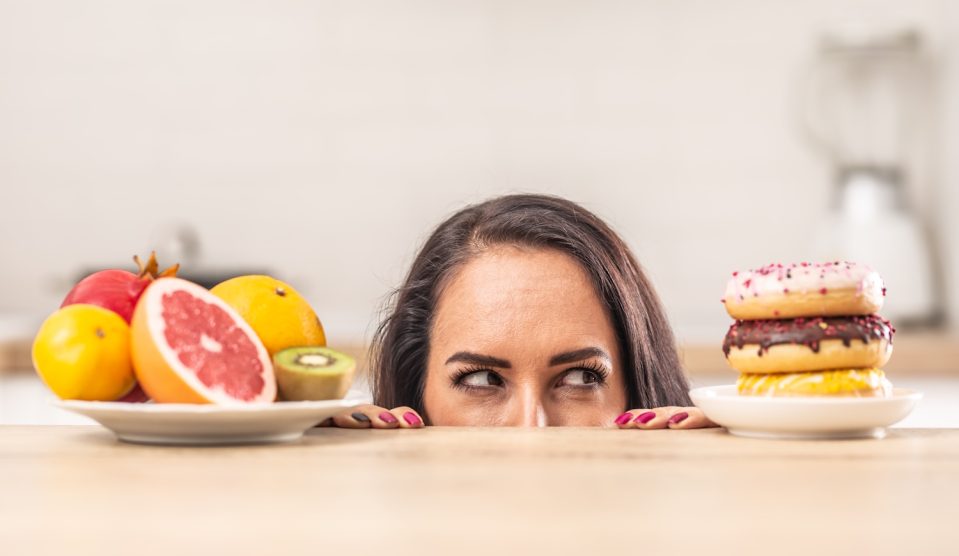 fogyni akaró nő nézi a fánkokat miközben van mellette egy tál gyümölcs