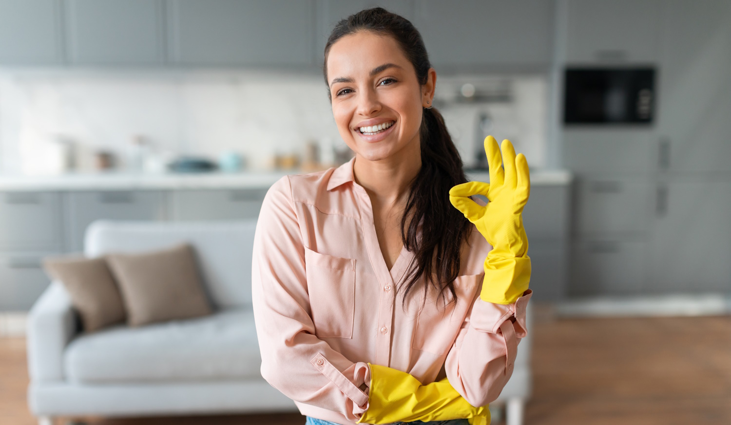 Pasztellszínű ingben és farmerben, sárga tisztítókesztyűben, a kamerába mosolygó, ragyogó nő készen áll arra, hogy megkezdje tavaszi nagytakarítását egy rendezett otthonban.