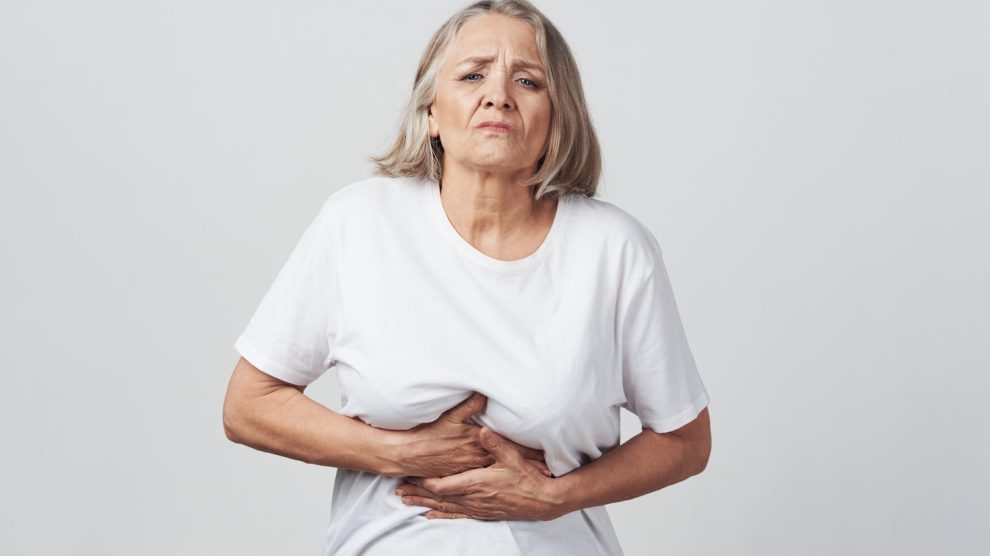 idős nő fehér pólóban a hasát fogja, mert fáj neki