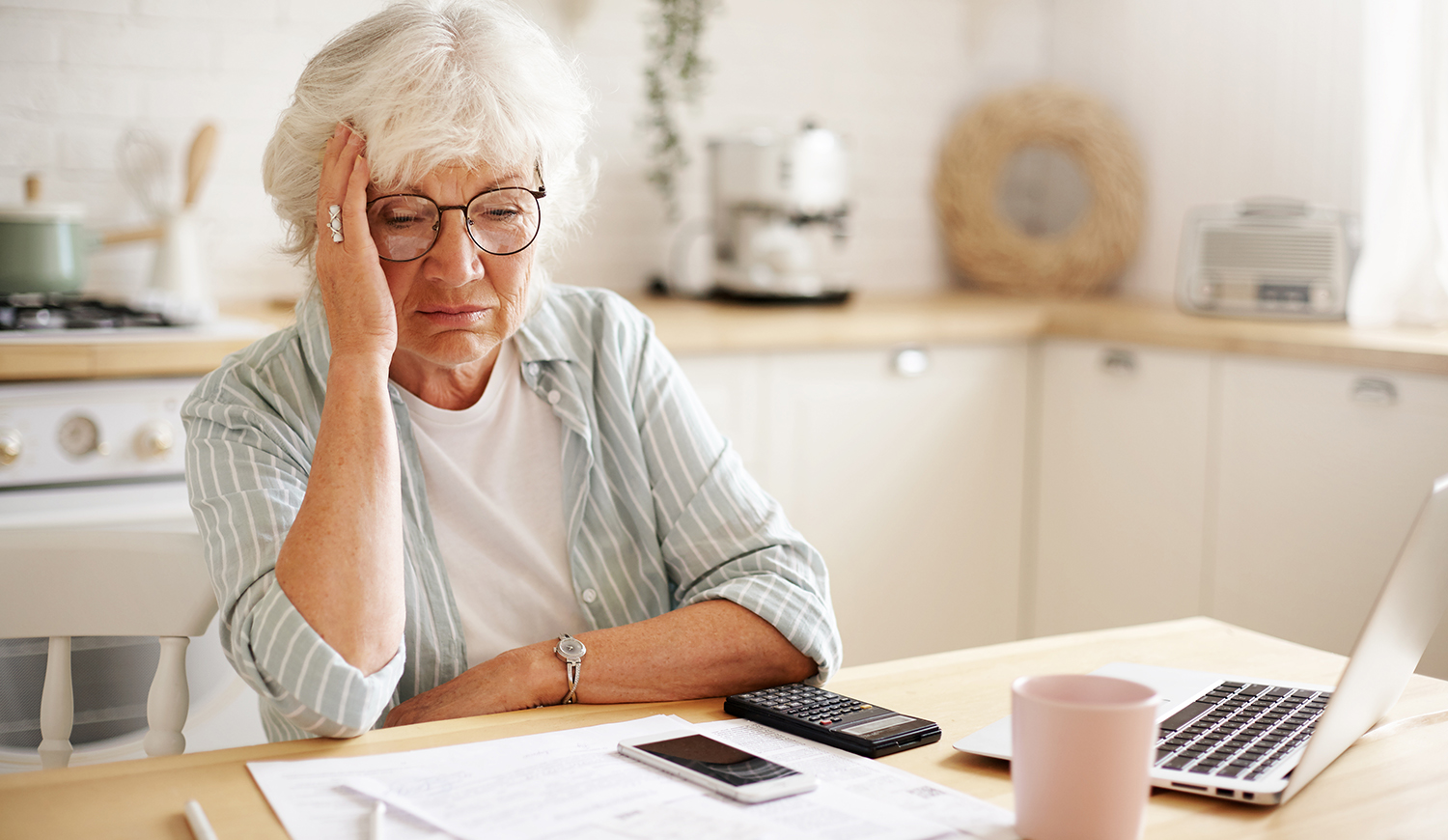 Szomorú nyugdíjas nő, aki a kezét az arcán tartja, családi költségvetést számolgatva ül a konyhában a laptop előtt