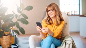 boldog középkorú nő a mobiltelefonját nyomkodja a nappaliban