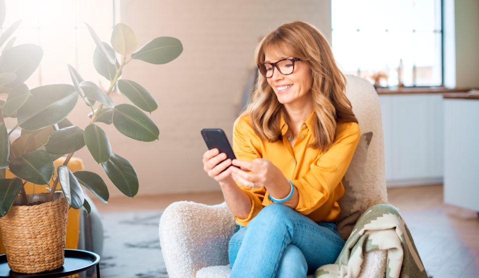 boldog középkorú nő a mobiltelefonját nyomkodja a nappaliban