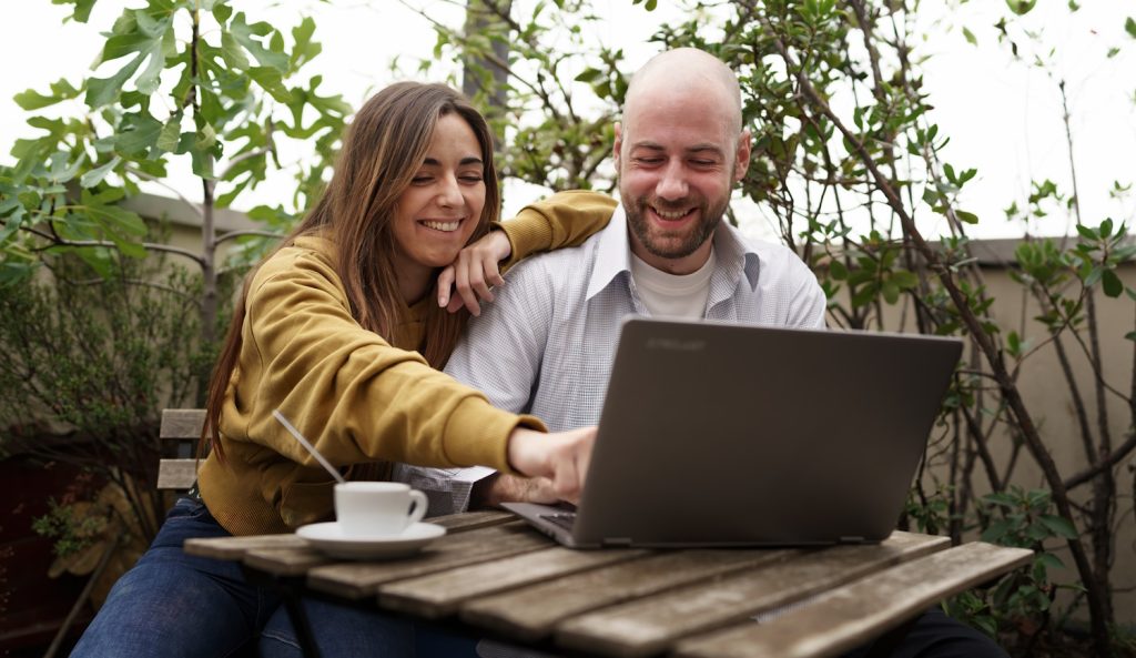 Fiatal pár kávézik és laptopot néz egy szabadtéri kávézóban – egy kopasz férfi és egy hosszú hajú nő nevetgél, miközben együtt böngészik a laptopot.