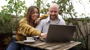 Fiatal pár kávézik és laptopot néz egy szabadtéri kávézóban – egy kopasz férfi és egy hosszú hajú nő nevetgél, miközben együtt böngészik a laptopot.