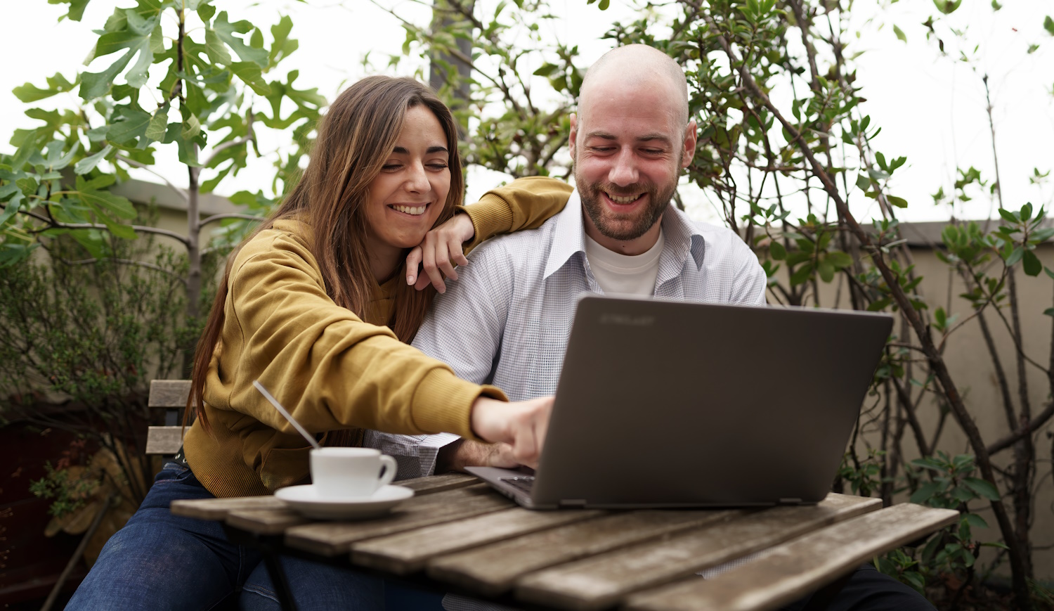 Fiatal pár kávézik és laptopot néz egy szabadtéri kávézóban – egy kopasz férfi és egy hosszú hajú nő nevetgél, miközben együtt böngészik a laptopot.