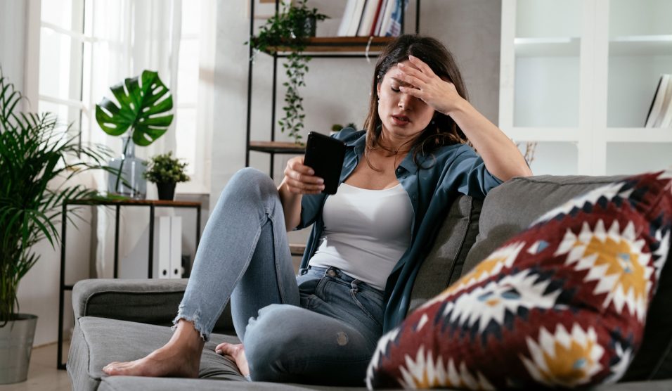 szomorú nő ül a kanapén, a telefonját nyomkodja