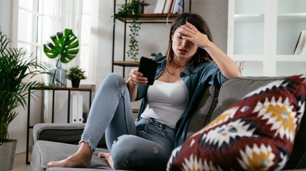 szomorú nő ül a kanapén, a telefonját nyomkodja