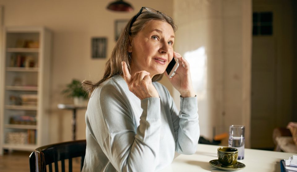 középkorú nő telefonál otthon a konyhaasztalnál