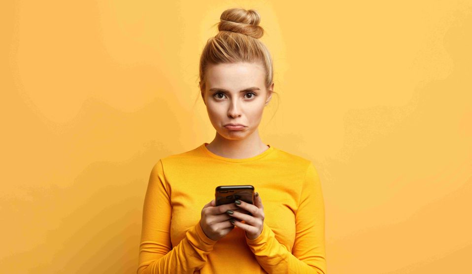 Nő sárga háttér előtt áll a telefonjával