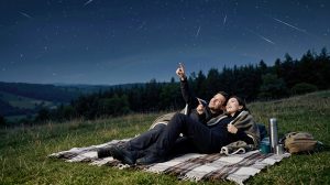 Egy fiatal pár egy kockás takarón fekszik egy füves domboldalon éjszaka, és a Perseidák meteorraj alatt figyelik a csillagokat.