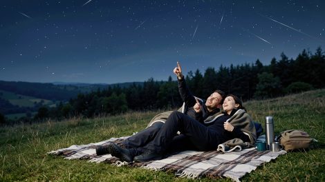 Egy fiatal pár egy kockás takarón fekszik egy füves domboldalon éjszaka, és a Perseidák meteorraj alatt figyelik a csillagokat.