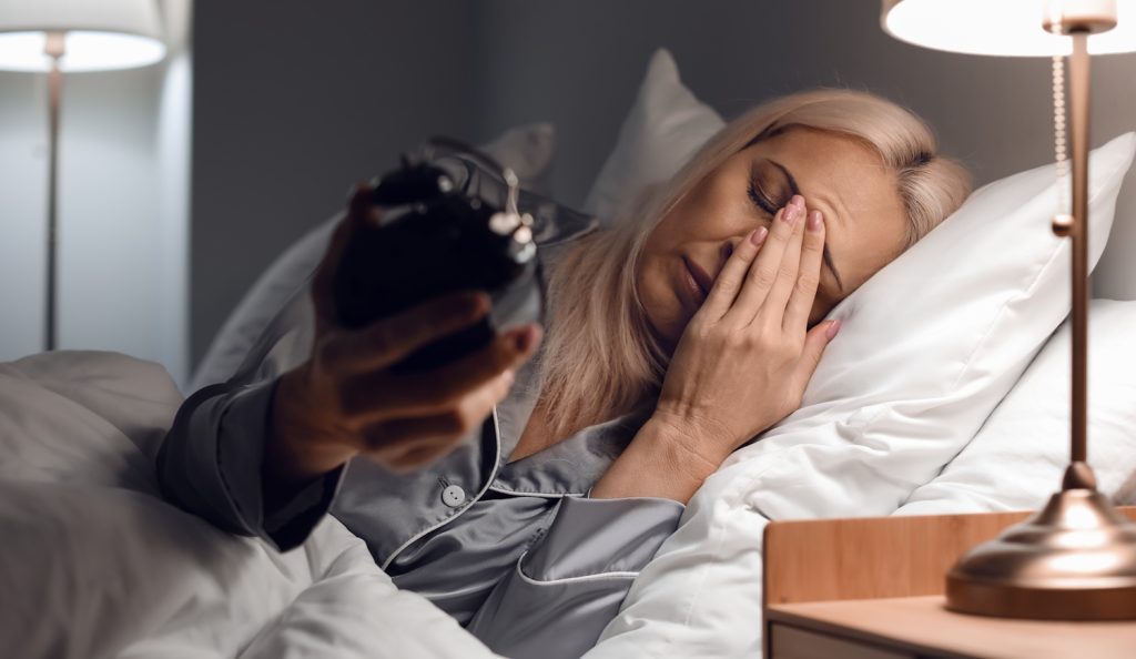 Stressed mature woman with alarm clock lying in bed at night | Meglepetés stresszes 60-as nő felébred az éjszaka közepén és az órájára néz