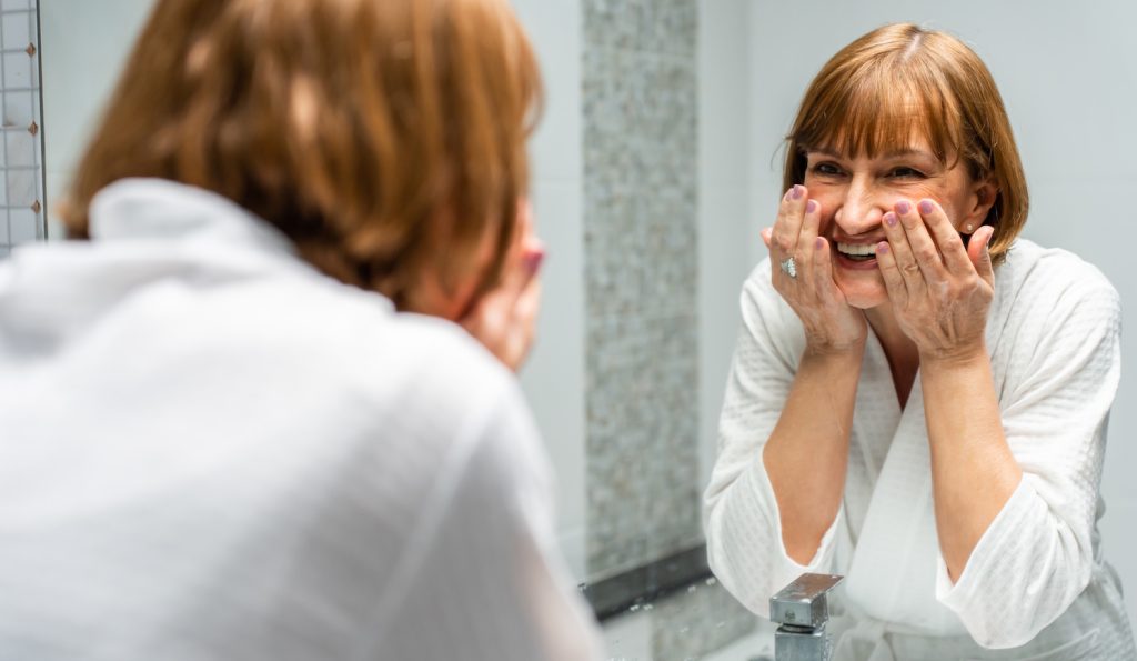 60+-os szép bőrű nő arcot mos a fürdőszobában