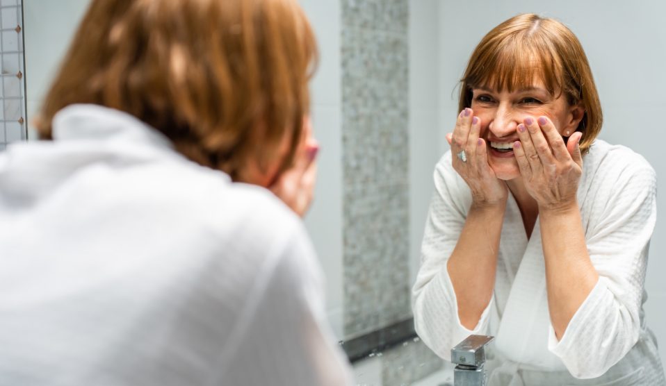 60+-os szép bőrű nő arcot mos a fürdőszobában