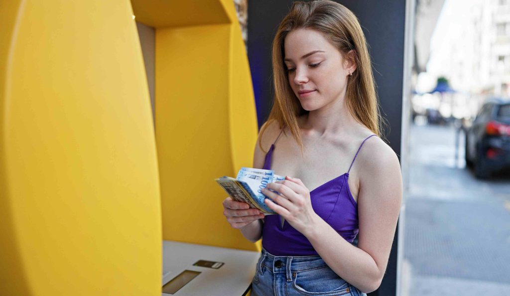 Fiatal nő bankjegyeket tart a kezében egy utcai bankautomatánál