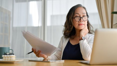 Papírokkal a kezében nézi egy nő a laptopját